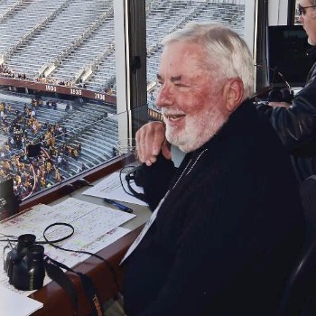 A man sits in a sportscaster booth. The view outside of the window show stadium seating half filled with people. 