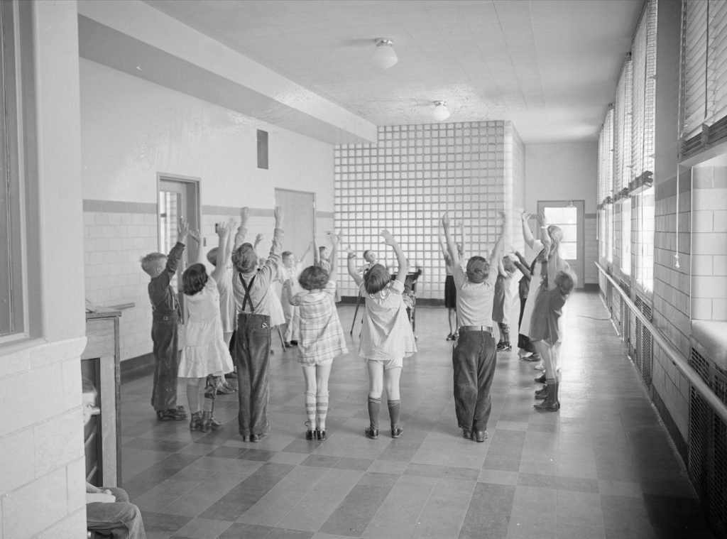 A teacher leads young children in stretching exercises in a large room. The children stand facing each other in a circle and have their hands raised over their heads. 