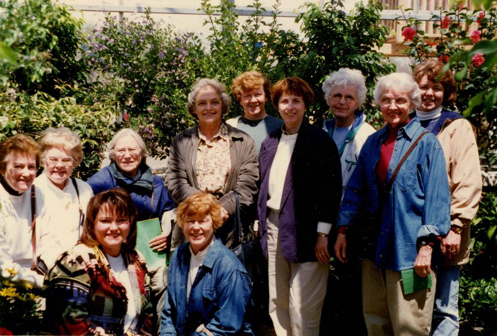 A group of women stand together in front of a backdrop of plants and greenery. 