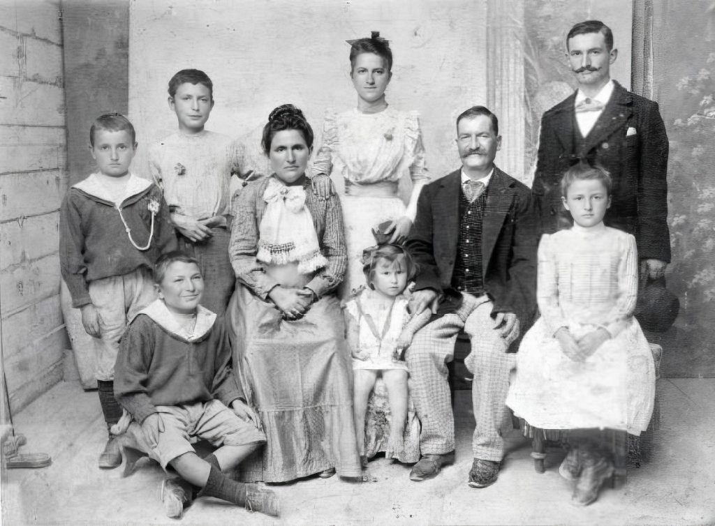 A family photo with a mother and father seated in the middle of their children. The children consist of 4 boys and 3 girls ranging from toddler to young adult. 