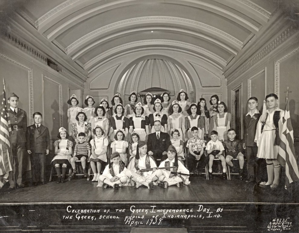 A large group of children pose on a stage for a group photograph. Several children are wearing traditional Greek clothing including vests and hats with long tassels on the point.