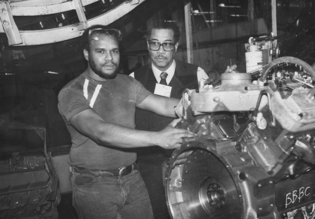 Two men stand behind a large engine in a factory. 