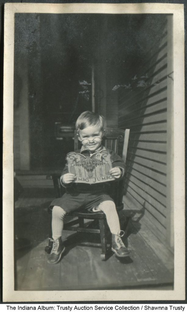 A child sits in a small chair on a porch and is reading a book. 