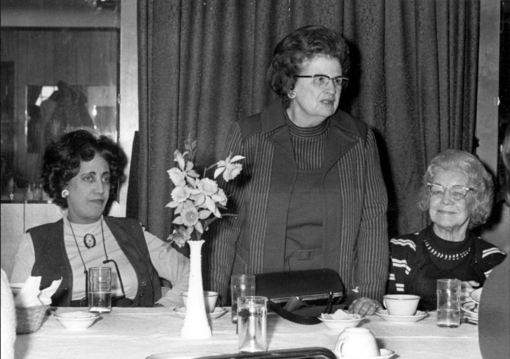 Three women are behind a table. The middle woman is standing and the other two women are sitting. 