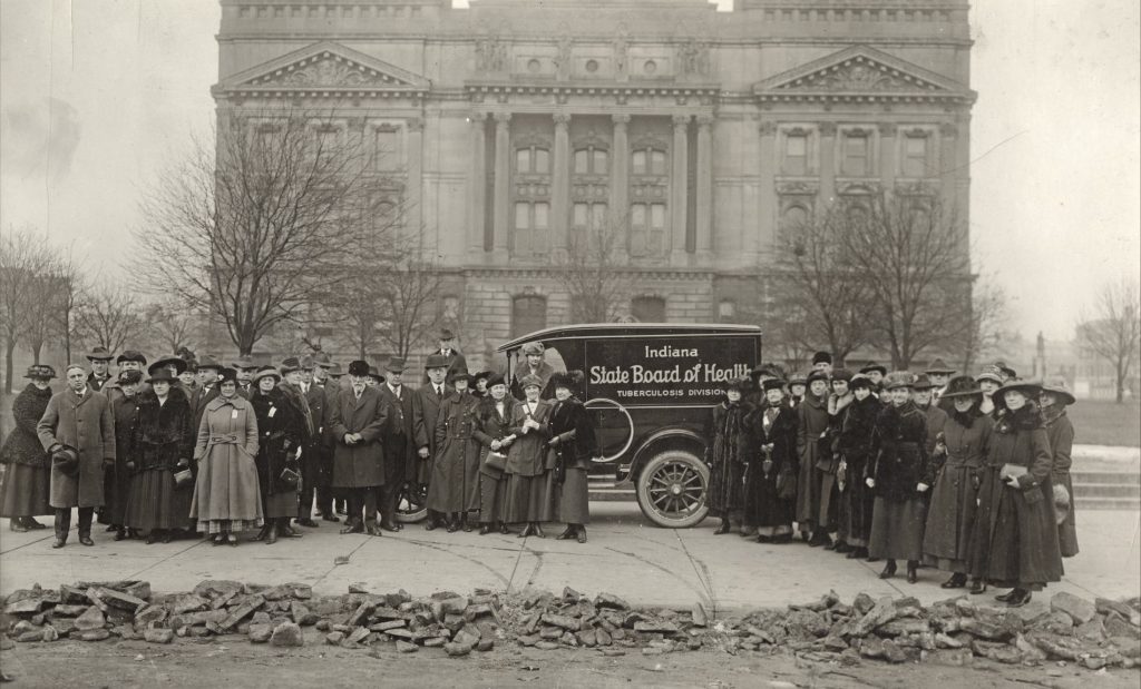A large group of people stand on either side of a car. The car has text that says "Indiana State Board of Health."