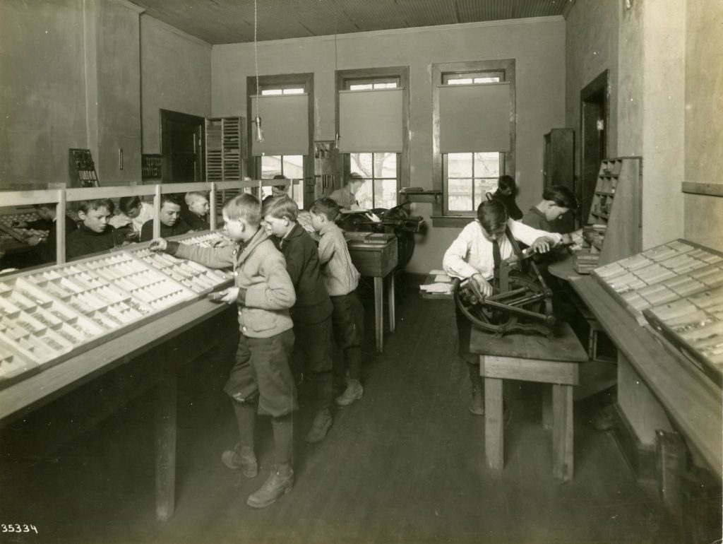 	 Classroom of boys working on various printing tasks. Some are standing at trays setting type, while others are working printing presses.