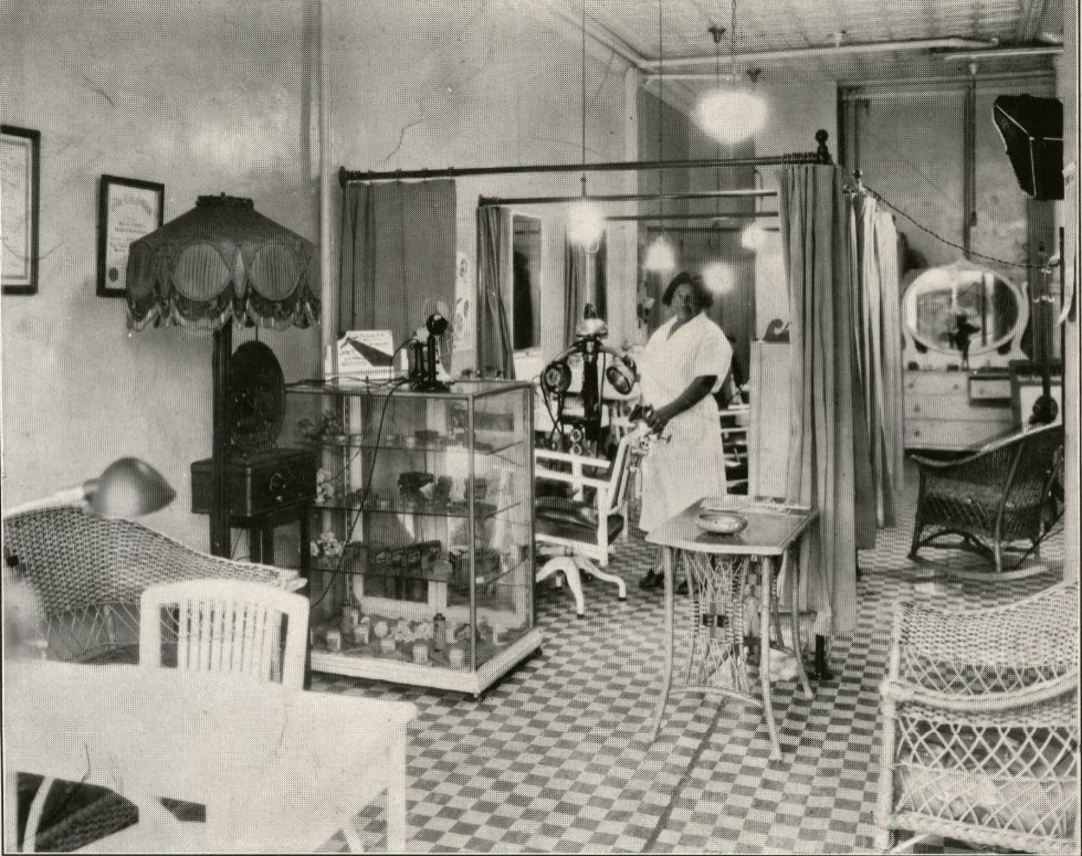 The interior of a Madam C.J. Walker Beauty Salon. The waiting area, manicure station and display case can be seen at the front. A salon employee stands in one of the stations next to a lamp.