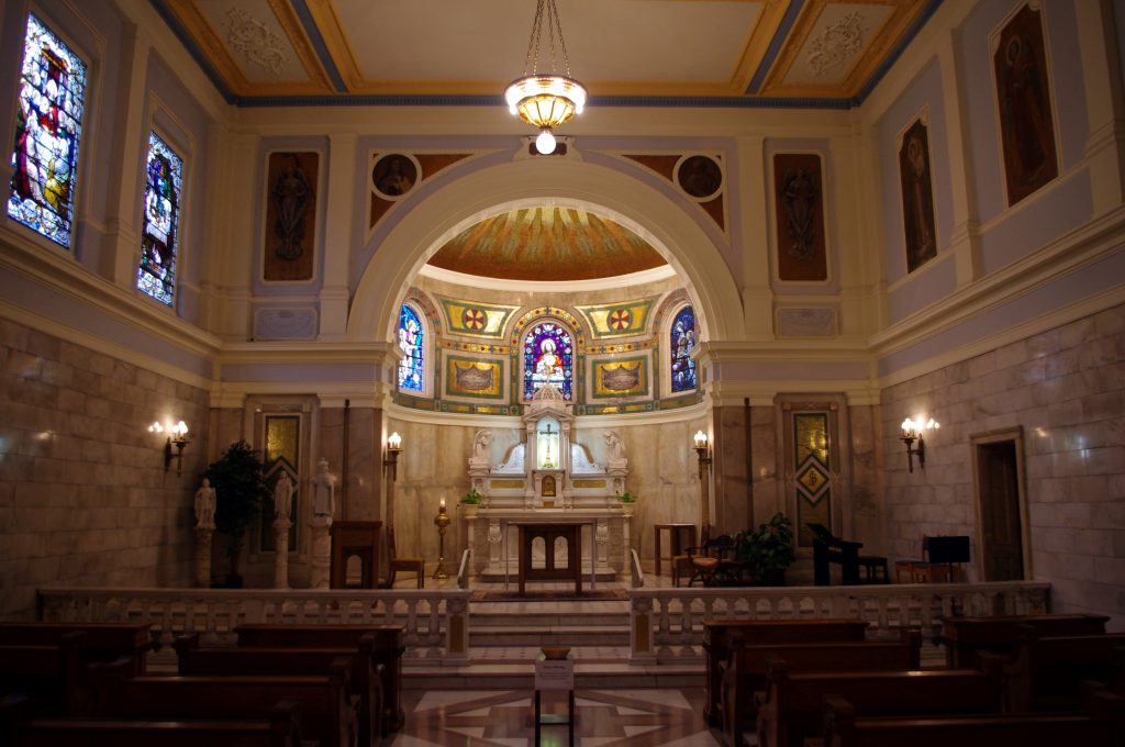 Interior of a chapel showing the sanctuary. The space is decorated with statues, stained glass, and an archway that leads to a domed ceiling space.