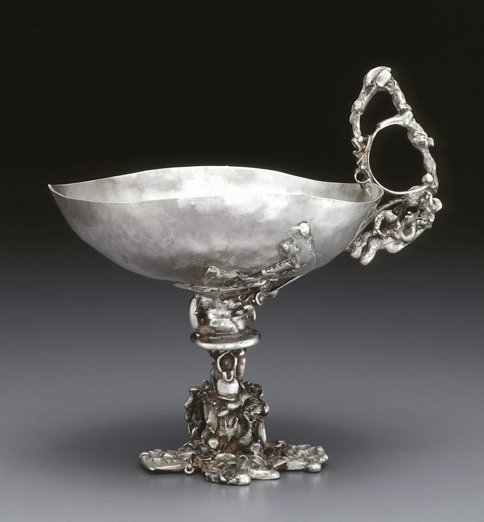 Silver metal chalice with an ornate handle on one side and an ornate stem.