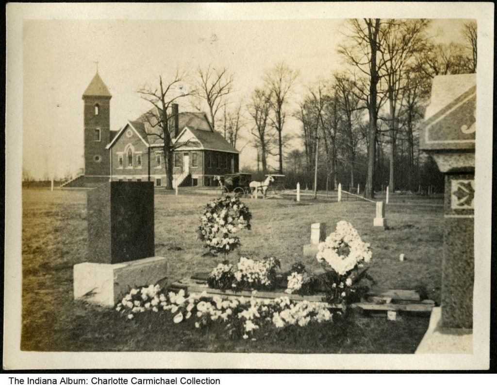 A granite headstone and flower-covered grave are in the foreground with a brick church in the background.