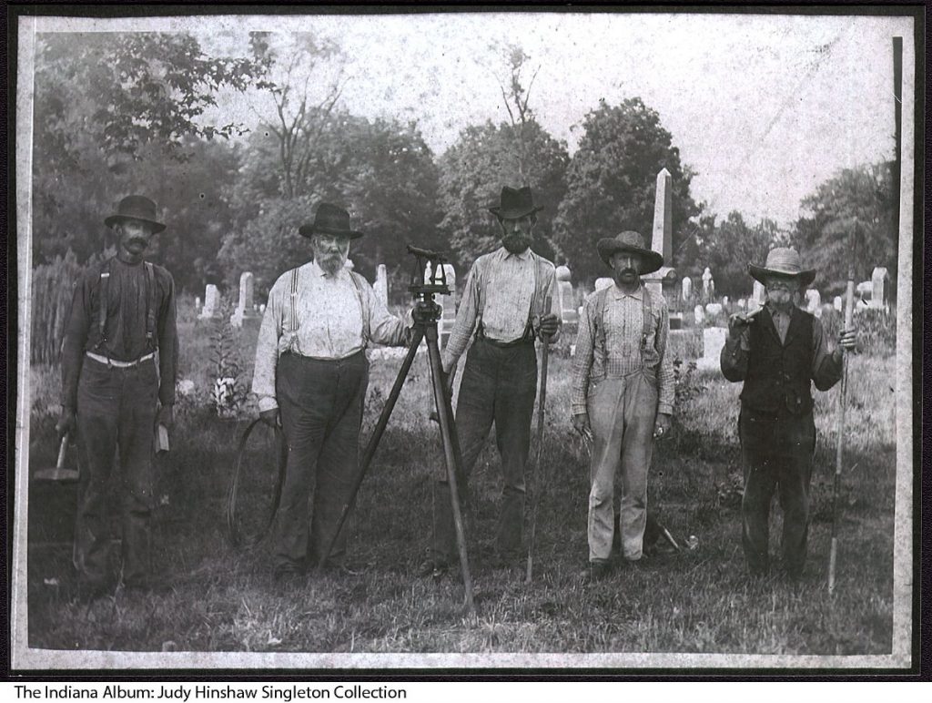 A row of five men with various tools stand in front of a cemetery.