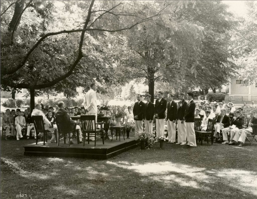 A group of people are gathered outside under trees. Several young men are lined up in front of a stage upon which a man is standing and talking in front of a group of people seated in chairs.