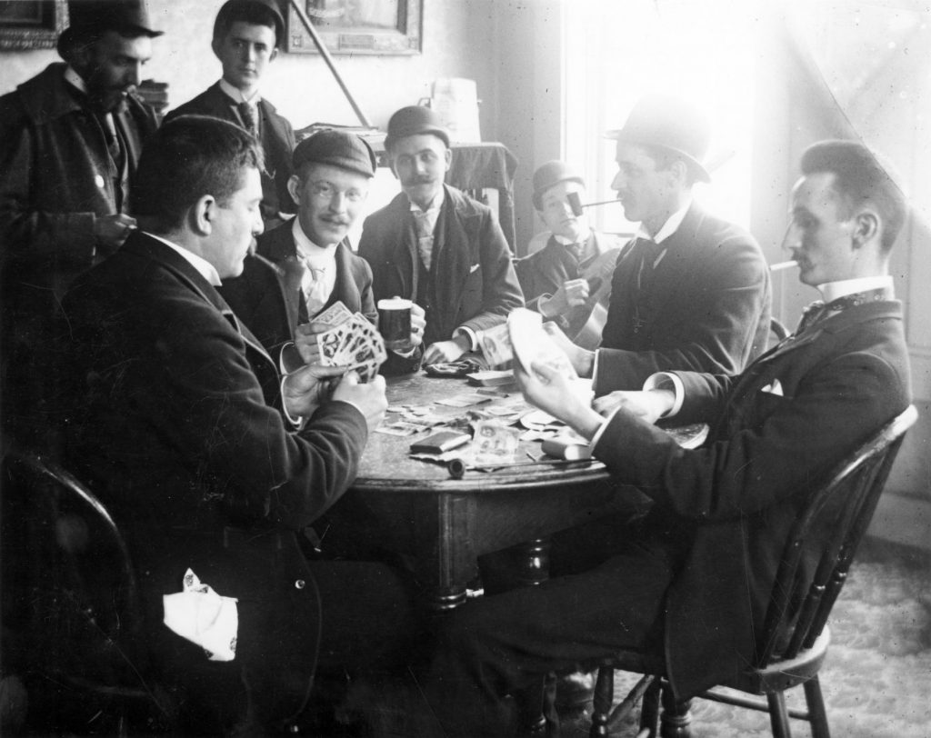 Four men, smoking and playing cards, are joined by several onlookers, one of whom strums a guitar.  