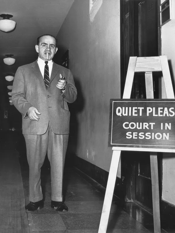 A man stands in a hallway and is about to light the cigarette in his mouth. A sign reads "Quiet please. Court in session."