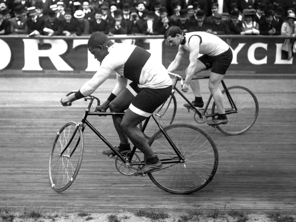 Two men are racing on bicycles. Major Taylor is slightly ahead of the other cyclist.