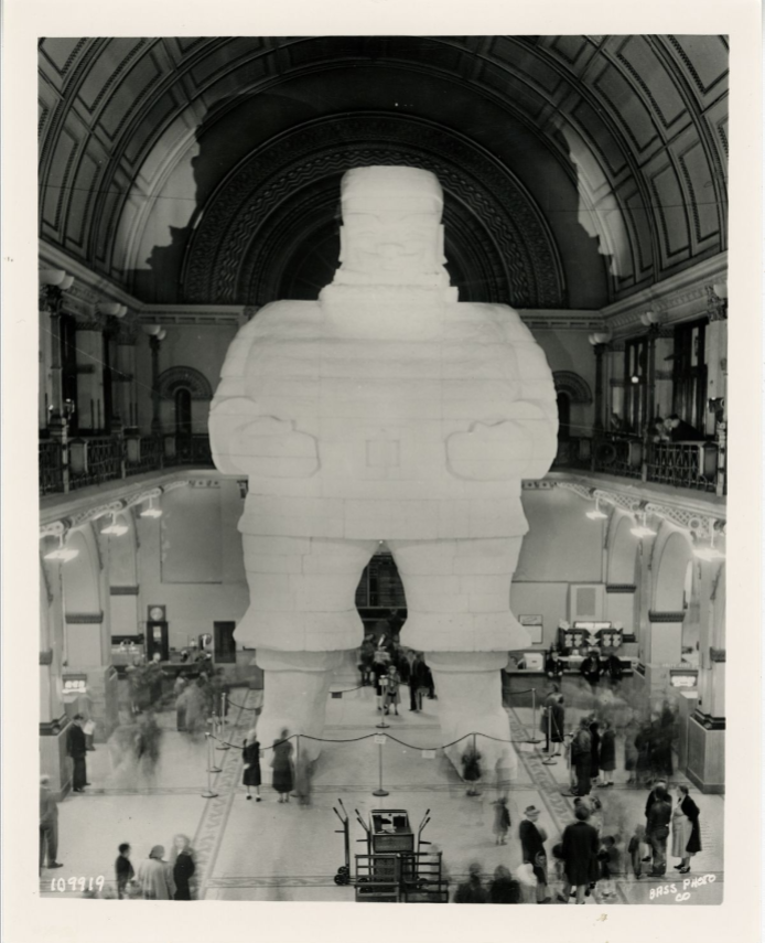 A giant styrofoam statue depicting Santa Clause stands in the middle of a large, multistory room.