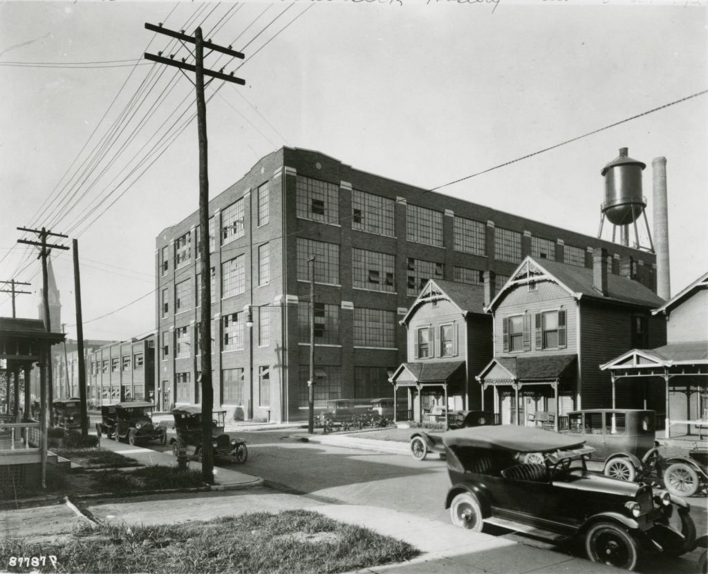 View across a street towards the Real Silk Hosiery Company. Some of the windows on the building are open. A water tower is on the roof.  The factory is located in a neighborhood with two-story wooden homes. Automobiles are parked along the street.