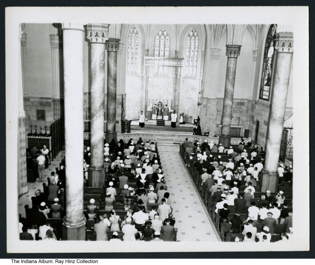 Interior of a church with people filling rows of pews. Clergy members are on a platform at the front of the room.