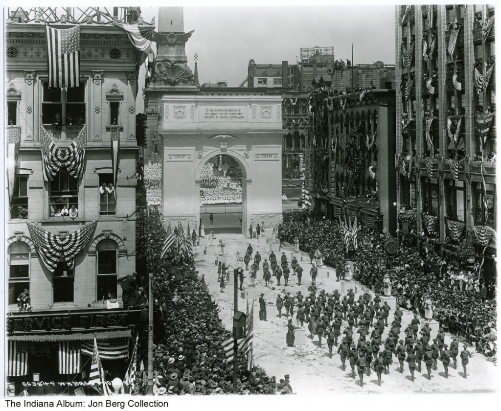 A large crowd is seen at a welcome home parade to celebrate the end of World War I. Headed north on Meridian Street is a military band marching past flower girls dressed in white standing on pedestals. At the entranced of Monument Circle is a large, temporary plaster arch with the dates 1917 and 1919 and the phrase "To The Returning Heroes Of The Great War We Extend This Welcome In Deepest Appreciation." 