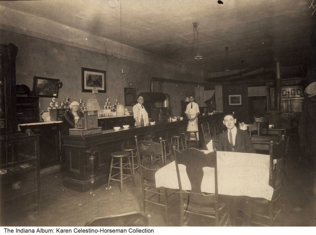 A lunch counter runs along the left wall with three staff members standing by. The rest of the tin-roofed room has tables and chairs.