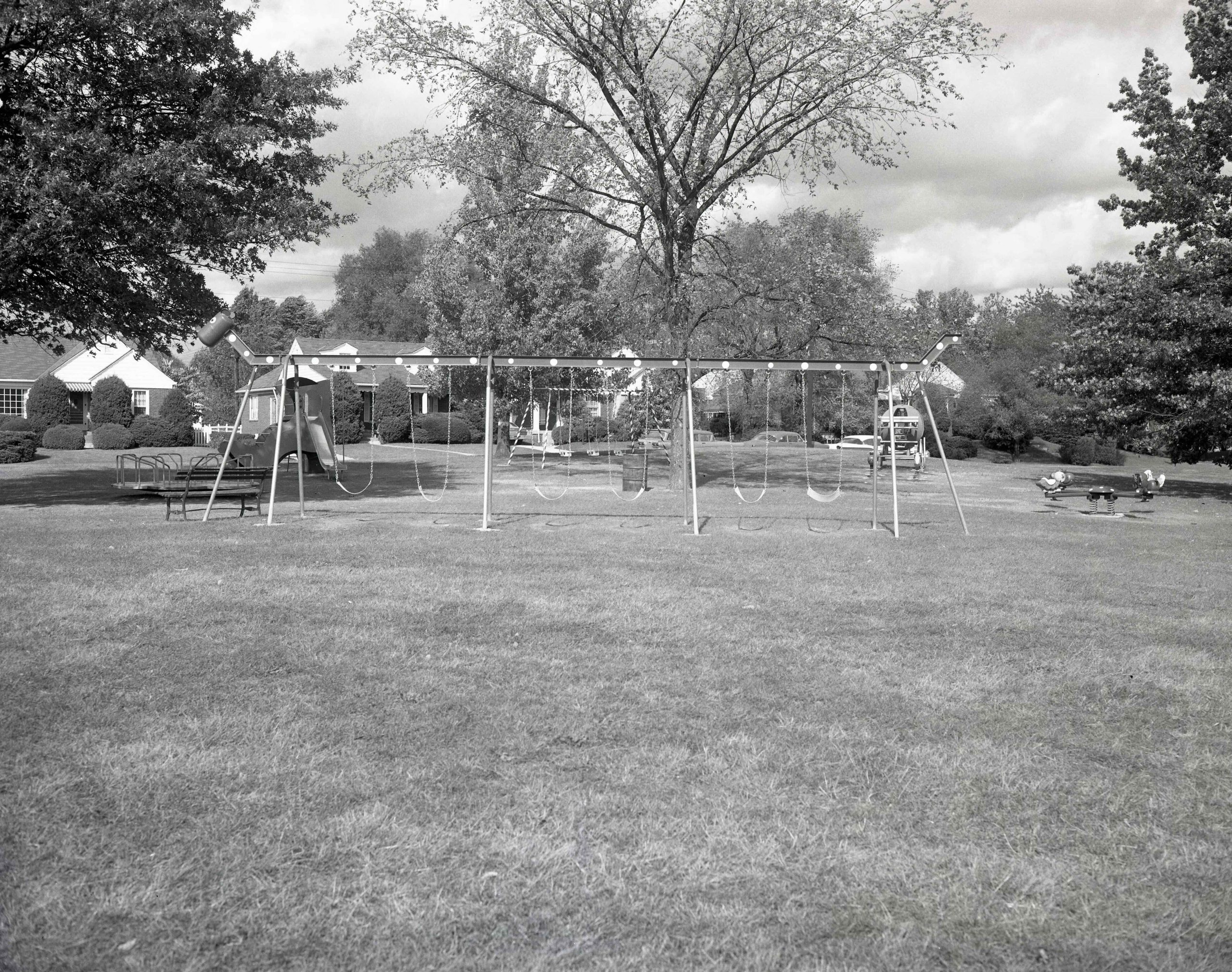 A park with two swing sets, a merry-g-round, and a slide. Small single family houses line the street behind the playground. 