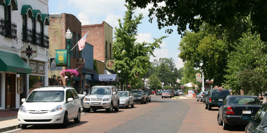A view down a paved road. Businesses line the road. Cars are parked along the sidewalk.