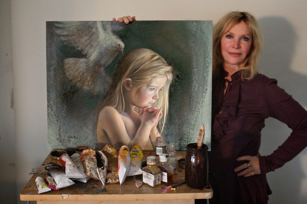 Nancy holds her painting of a young girl and a dove. Paint supplies are strewn on top of a table.