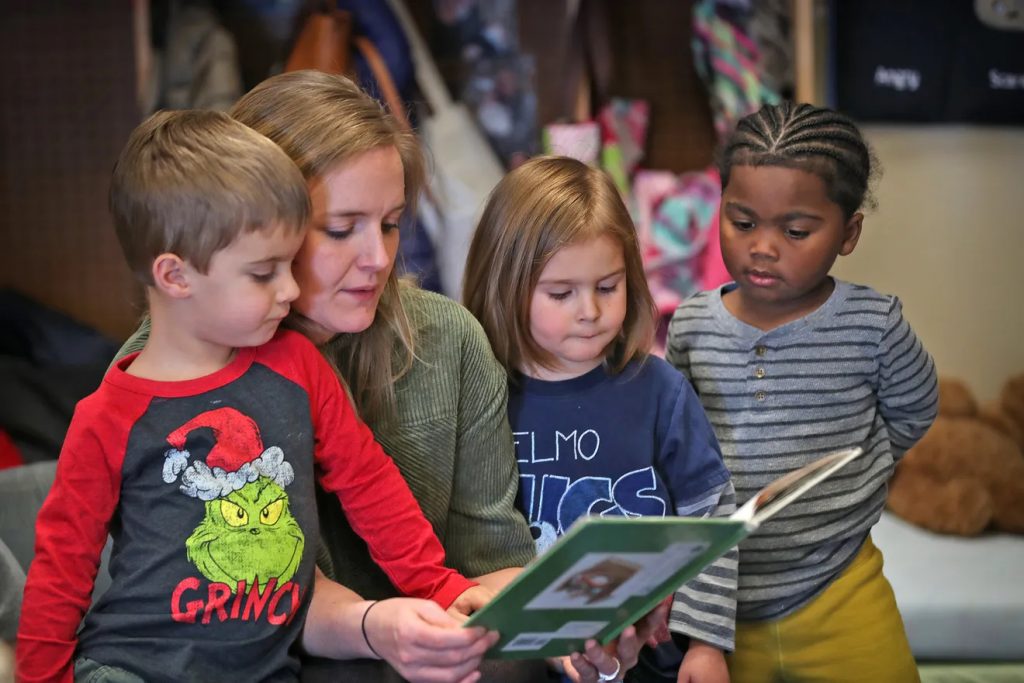 A woman holds a book and three small children gather around her.
