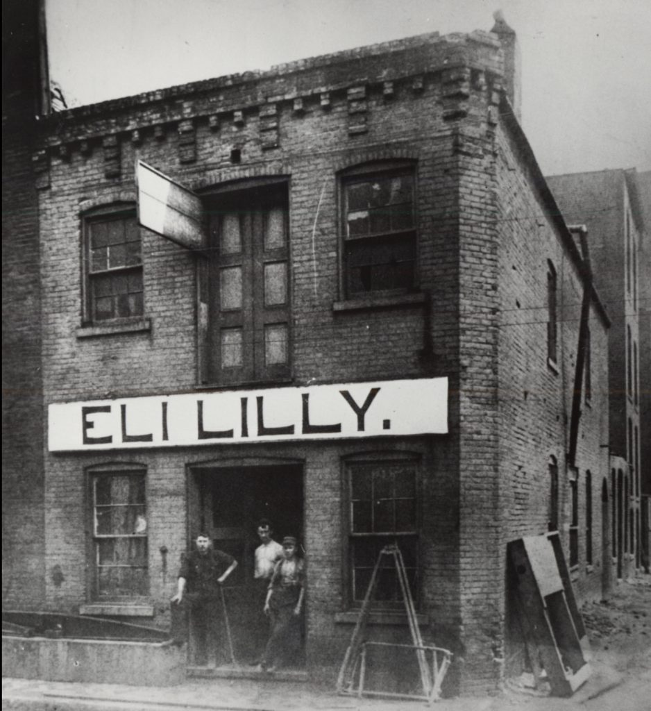 Exterior view of a two-story building with a large sign across the front that reads "Eli Lilly." Three men stand in the doorway of the building looking out.