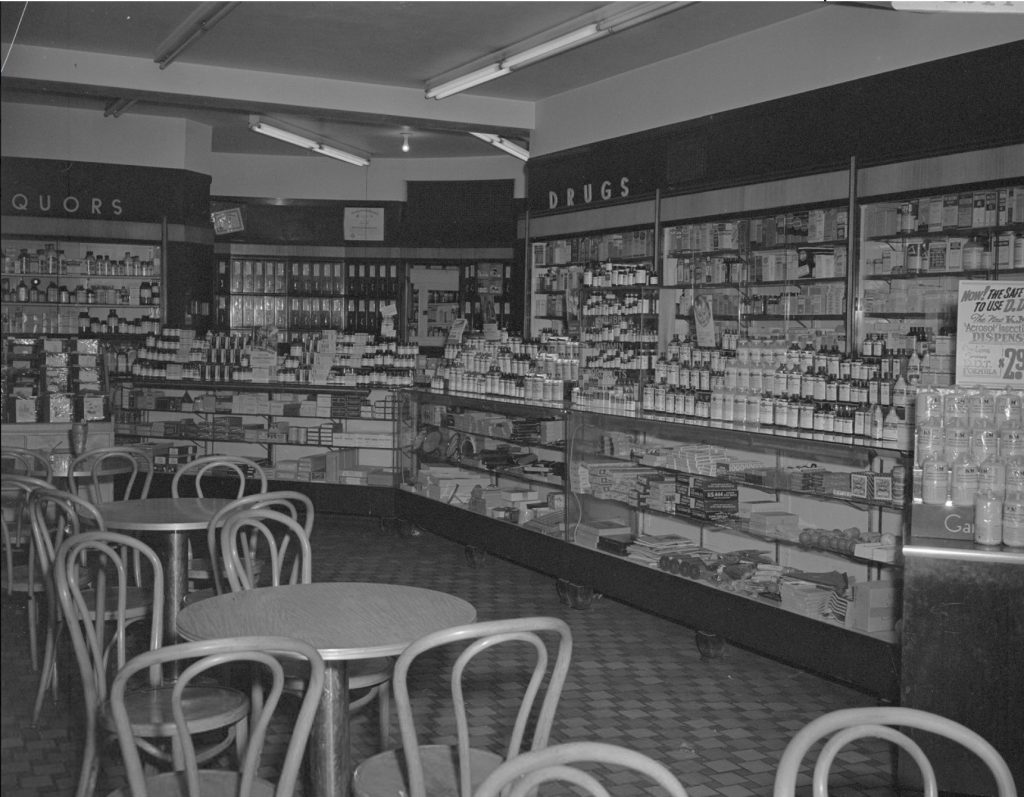 Interior view of a drugstore. Products line the walls and fill glass cases. Several small round tables with chairs fill the floor space. 