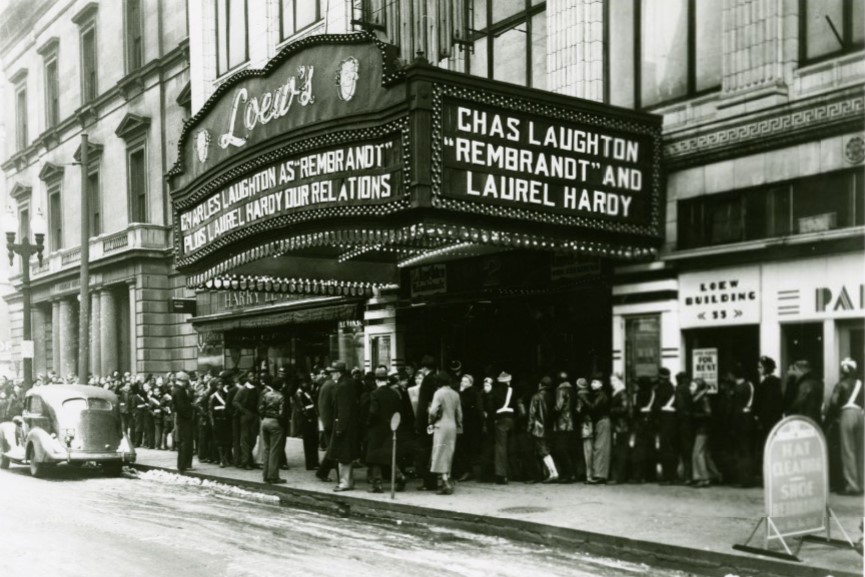 A large crowd stands outside the entrance of a theater. The theater marquee advertises two movies: "Our Relations" and "Rembrandt." 