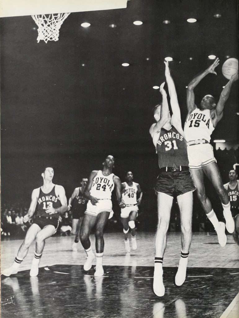 Two teams are running on a basketball court. In the foreground, one player is mid-air with the basketball in his hands while another jumps to block him. 