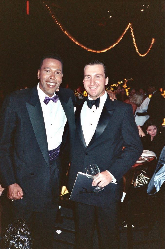 Two men in tuxedos stand next to each other. 