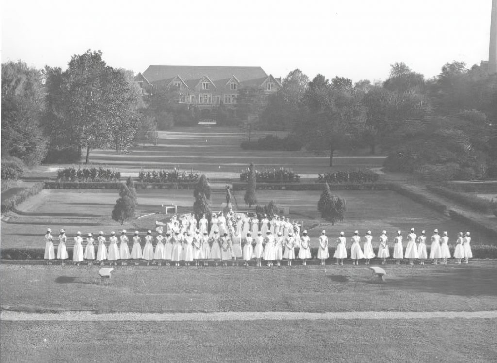 A large group of women in nursing uniforms stand in a formal garden. 