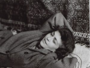 Jean Garrigue is wearing a coat and laying on a blanket with her hands behind her head.