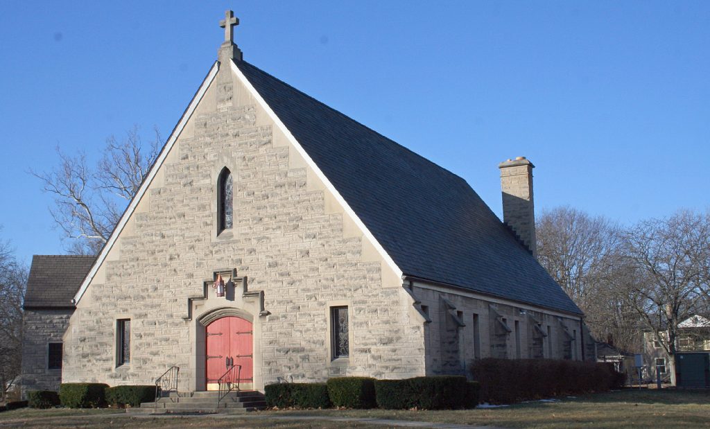 A church building of limestone with gable ends, a rose window over the chapel, and tongue and groove wood entrance doors.