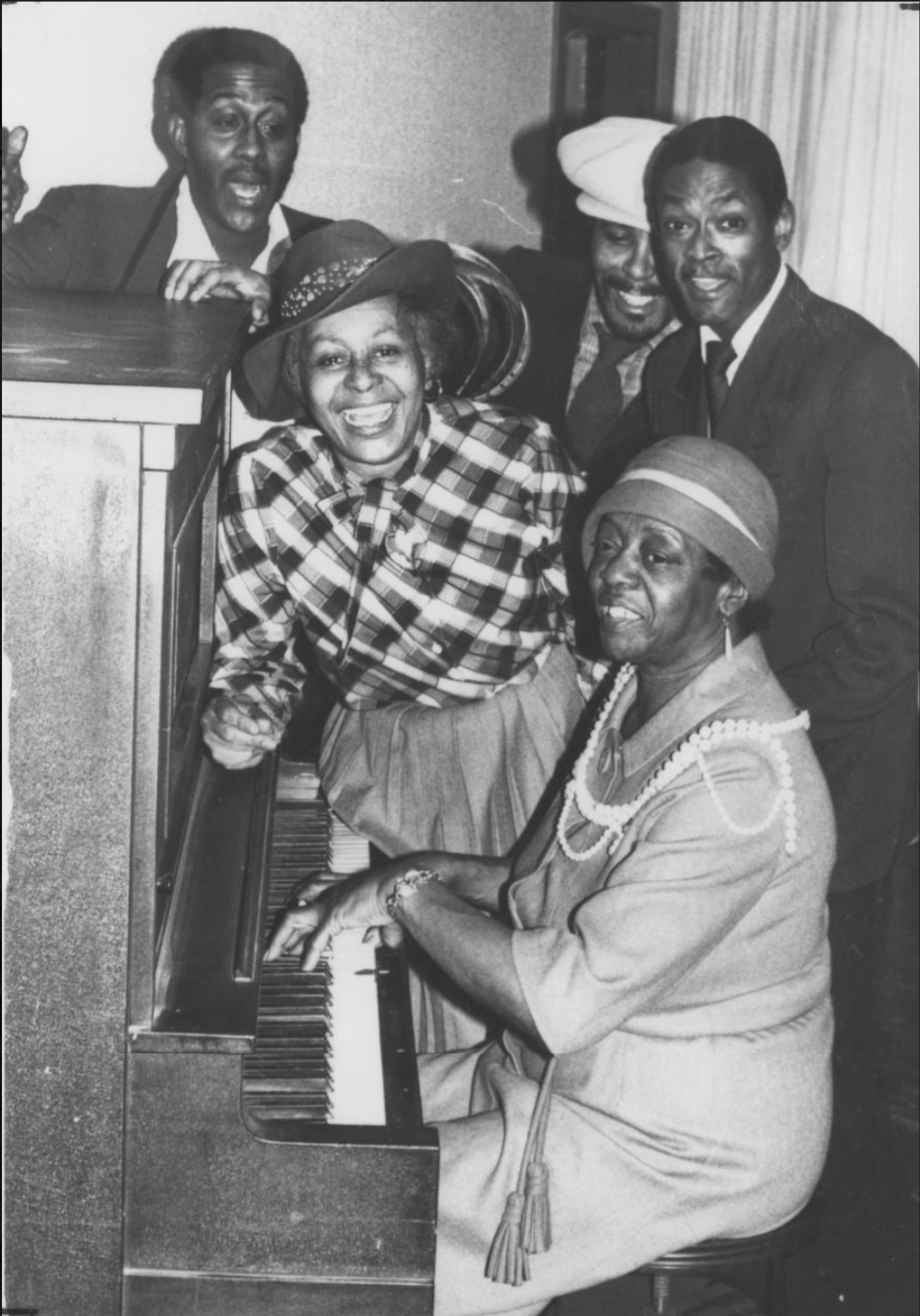Helen Whitelowe leans on a piano. Three people stand around her and one person sits at the piano.
