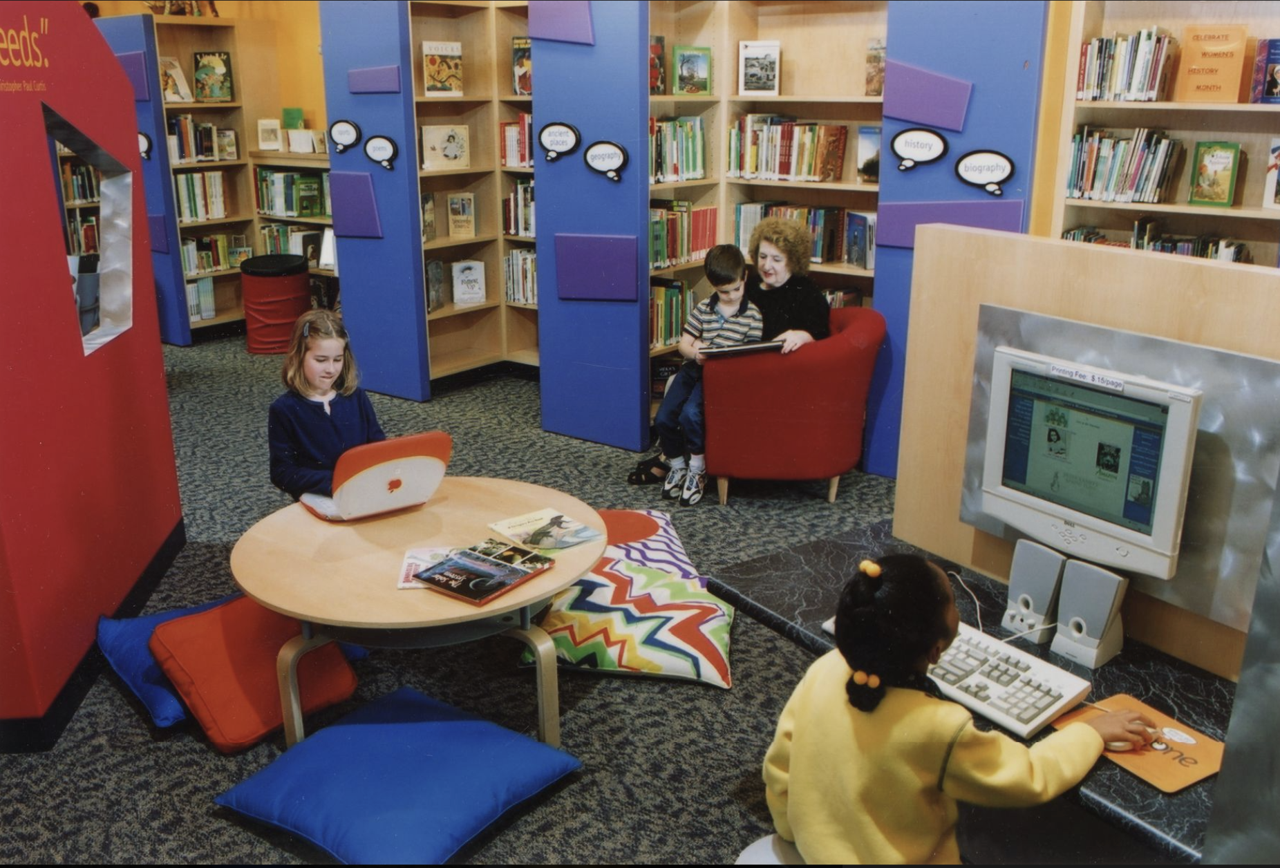 Two children use computers while another child sits in a chair with an adult showing them a book.