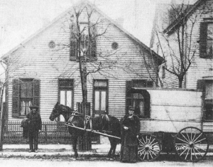 A man and woman stand in front of a small house with a horse and carriage.