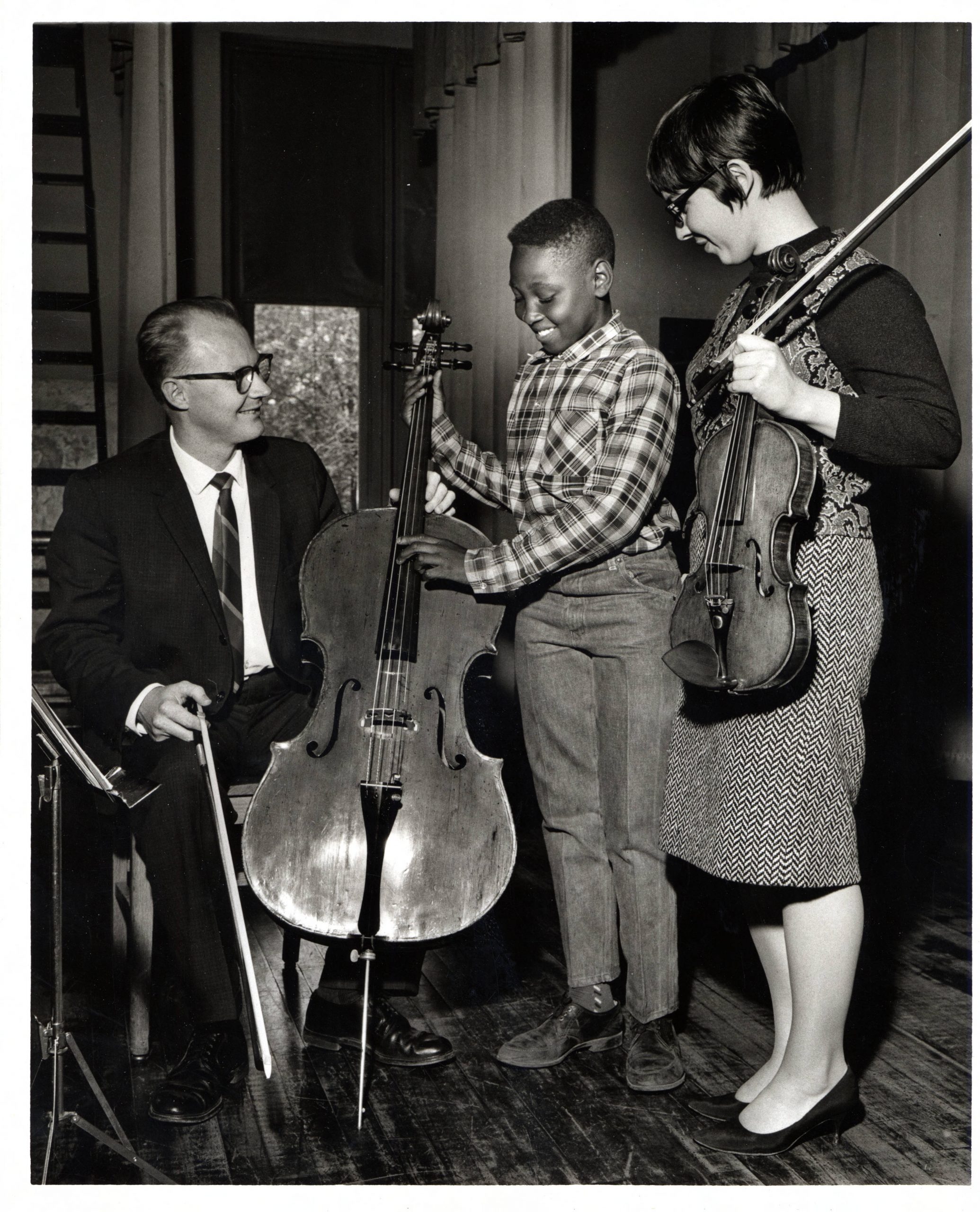 A man is seated while showing a child how to play a cello. A young woman looks on while holding a violin.