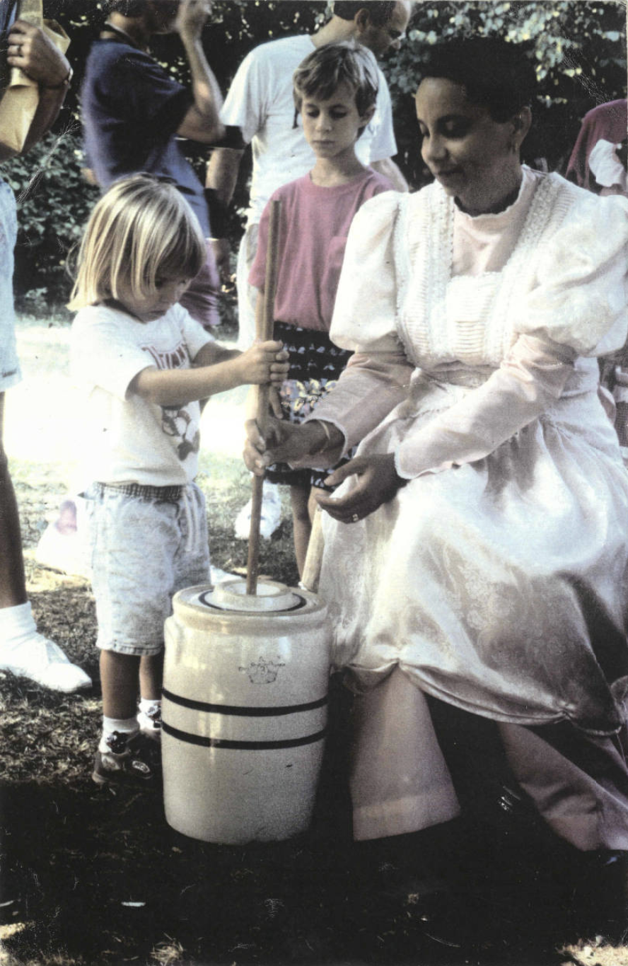 Freetown village costumed interpreter demonstrates to a child how to churn butter.