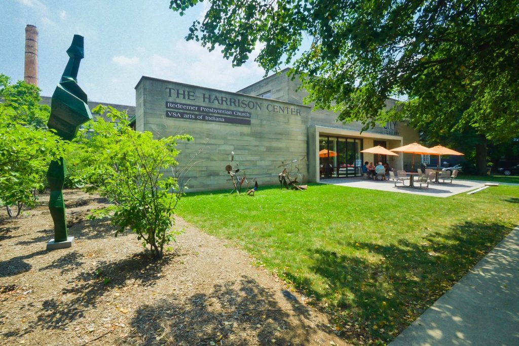 Exterior view of the Harrison Center building with abstract sculptures decorating the lawn and people sitting at tables on a patio.