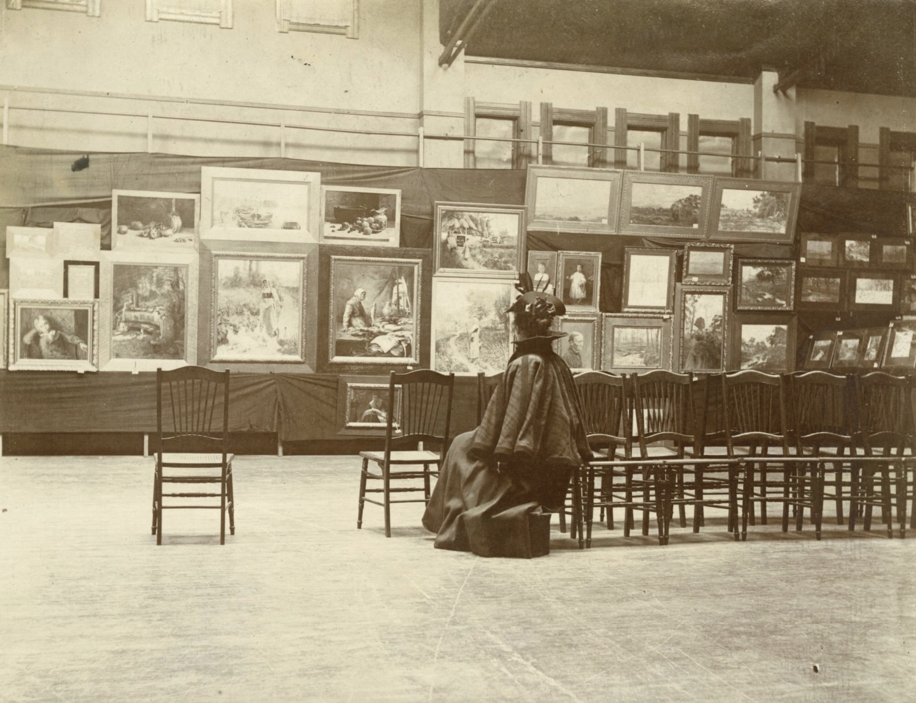 A woman sits alone in one of several chairs facing a wall with several paintings inside the Herron Art Institute Galleries.