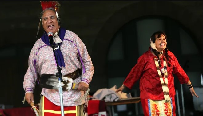 Two Native American people dressed in traditional clothing perform on a stage.