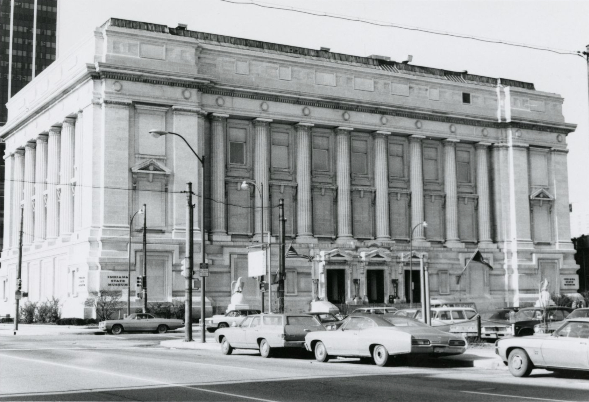 Exterior view of the front of a neoclassical style building. Cars are parked along the road next to the building.
