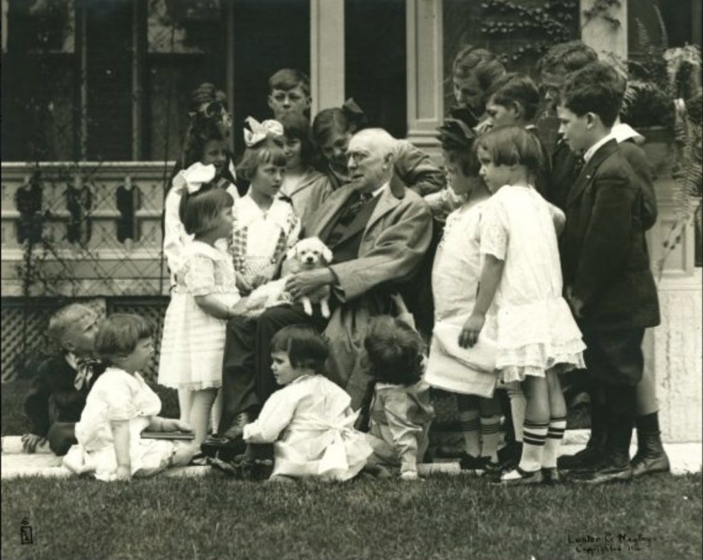 James Whitcomb Riley is seated outdoors with a small dog on his lap. Children stand around him.
