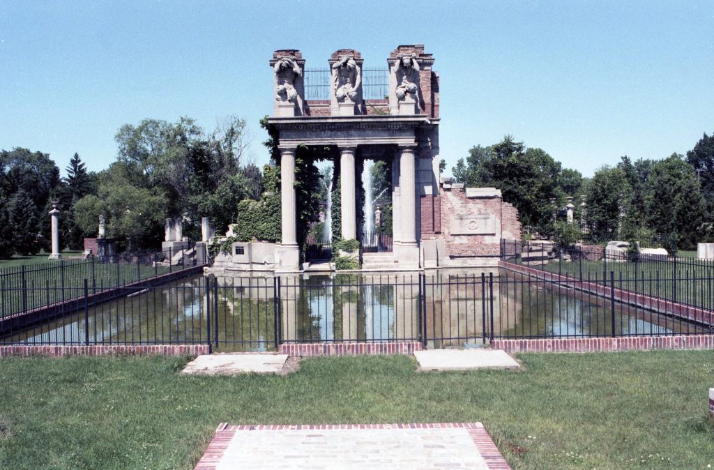 Art piece with three columns and three statues overlooking a pool of water.