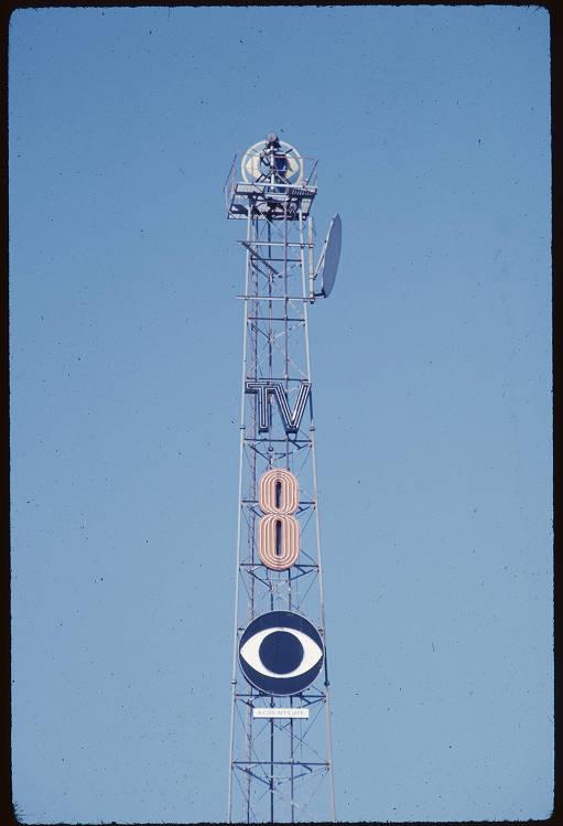 The top of a radio tower with large letters hung on it. The letters spell "TV 8" and under that is the eye-shaped logo for Channel 8.