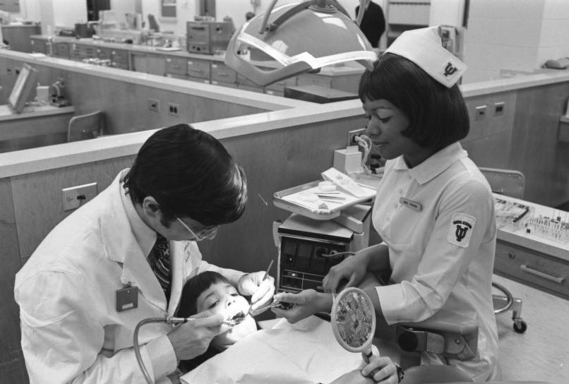 A child lies in a dentist chair while a man holds dental instruments and works on his mouth. A woman is handing the man an instument.