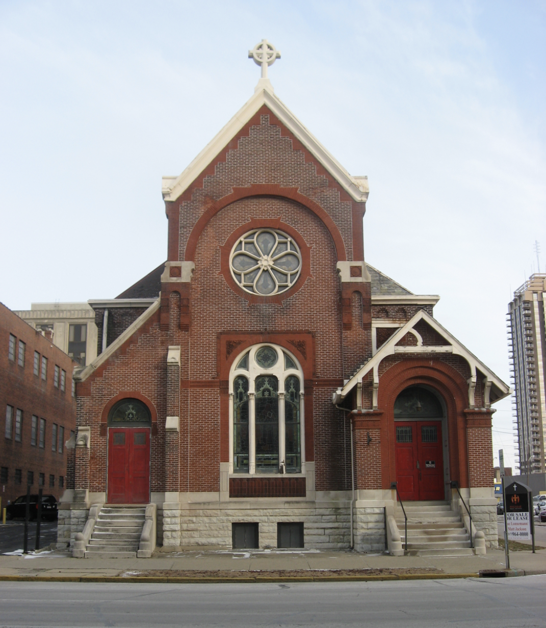 A brick church building with a large, round, central window above an arched window. The two entrances feature red doors.